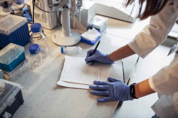A student taking notes in a laboratory notebook while surrounded by various lab equipment, demonstrating the importance of documentation in research.