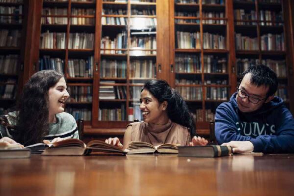 A group of three students studying in a cozy library room with wooden bookshelves filled with books. The students are smiling and appear to be enjoying a conversation while working together.