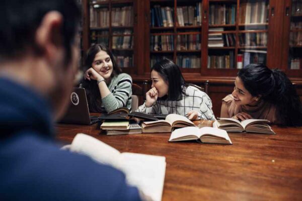 A group of young people in a sports setting, likely during a break from playing indoor soccer. They are standing by the side of the court near the bleachers, where a few spectators are seated. The scene is viewed through a mesh net, adding a dynamic effect to the image.