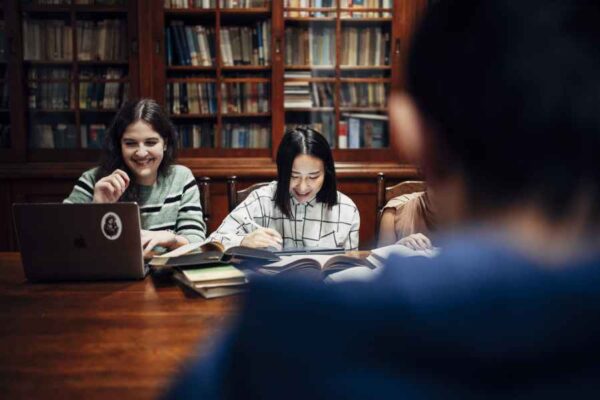 Three students studying together at a library table with books spread out in front of them. They are smiling and appear to be engaged in a light-hearted conversation. Behind them, wooden bookshelves filled with books create a scholarly atmosphere.