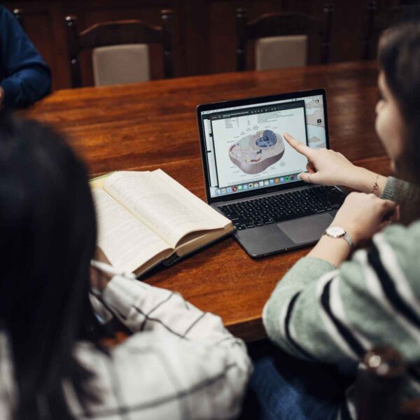 A different perspective of the library table with a focus on a laptop screen showing an academic diagram. A student is pointing at the screen while others listen, highlighting an educational discussion.