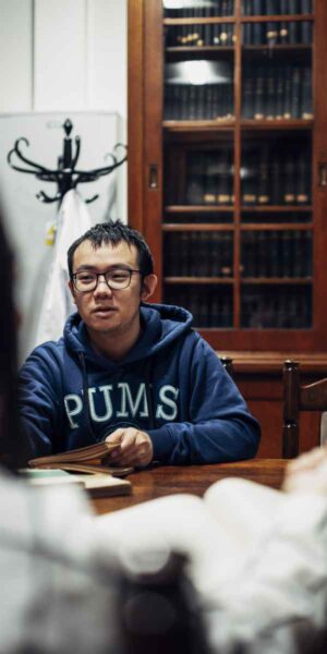 A closer shot of the male student in the "PUMS" hoodie, concentrating on the discussion. The background shows wooden bookshelves filled with books, reinforcing the academic environment.