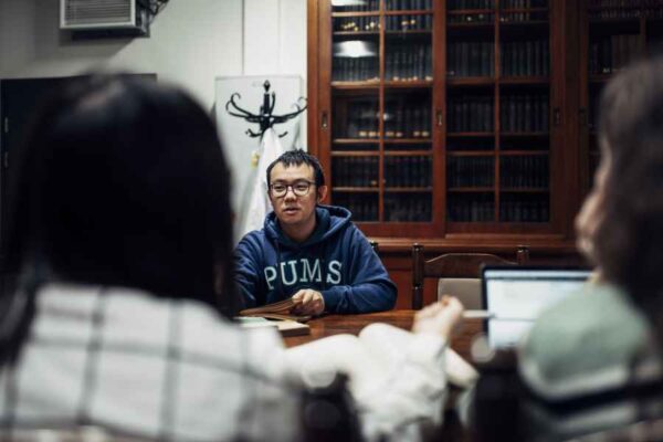 A male student, wearing a hoodie with "PUMS" written on it, leading a discussion at the library table. He appears to be explaining something to the others seated at the table.