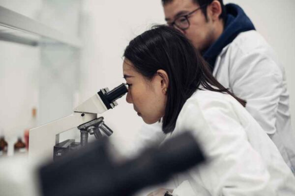 A female student closely examines something through a microscope, with the male student also observing. The laboratory setting includes scientific equipment, indicating an academic or research context.