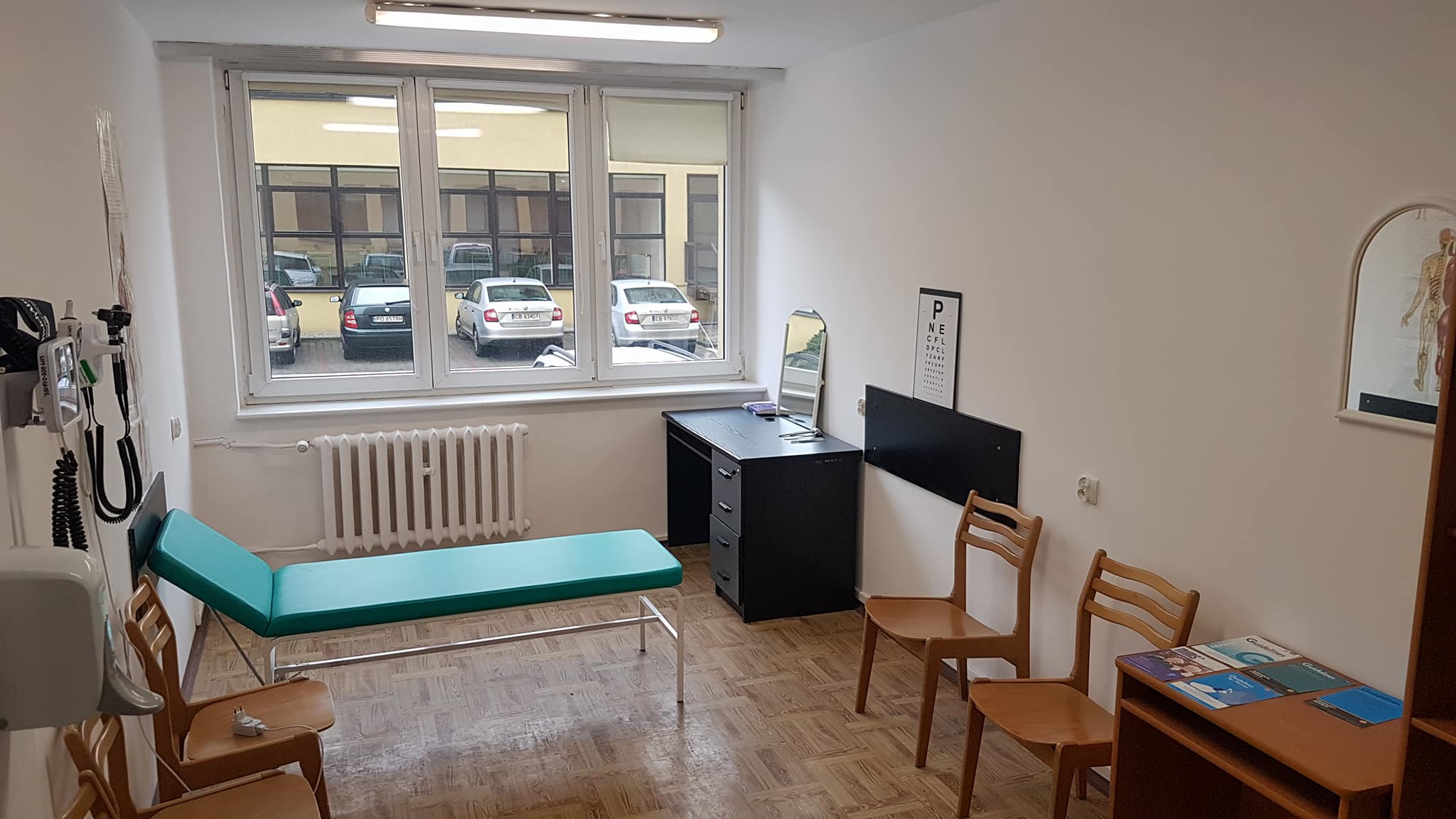 A small clinic room with basic medical furniture, including a green examination bed, chairs, and a window that shows parked cars outside. The room is clean and minimally decorated.