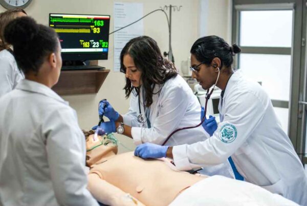 A medical team engaged in a hands-on simulation, providing care to a mannequin. The team members are dressed in white coats, focused on practicing medical procedures while monitoring the patient’s vitals on a screen.